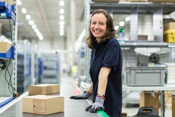 smiling young woman working in large distribution warehouse standing by conveyor belt.